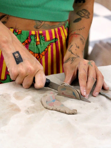 Person with tattoos using a tool on a piece of clay, creating ceramics by hand using manual techniques and nerikomi