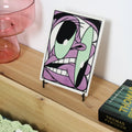 Tile with a Cartoon illustrated by Bráulio Amado on a tile stand next to a book of Thomas Pynchon on a wooden surface