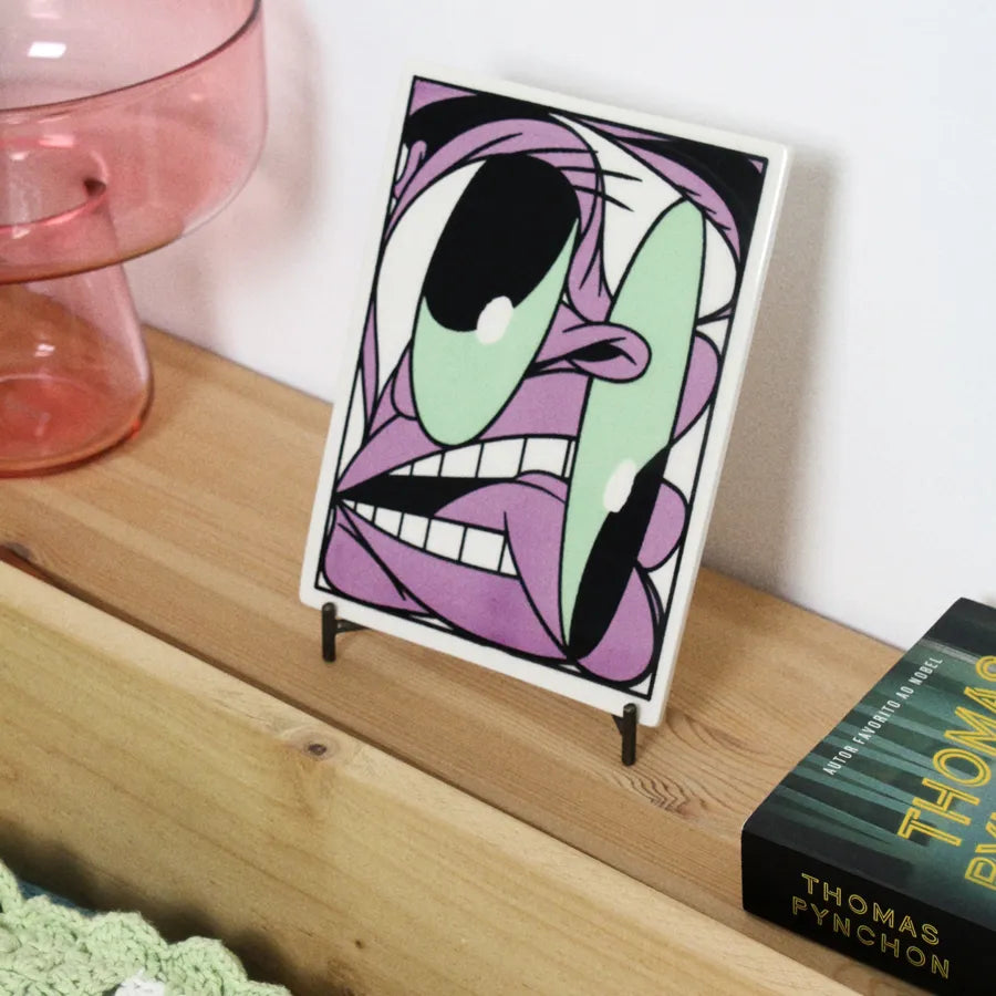 Tile with a Cartoon illustrated by Bráulio Amado on a tile stand next to a book of Thomas Pynchon on a wooden surface