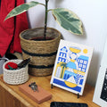 Small plant in a woven pot, crochet basket with yarn, keys, and a tile with an illustration on a wooden surface.