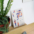 screen-printed tile with colorful illustrations of people on the snow on a wooden surface next to a potted plant.