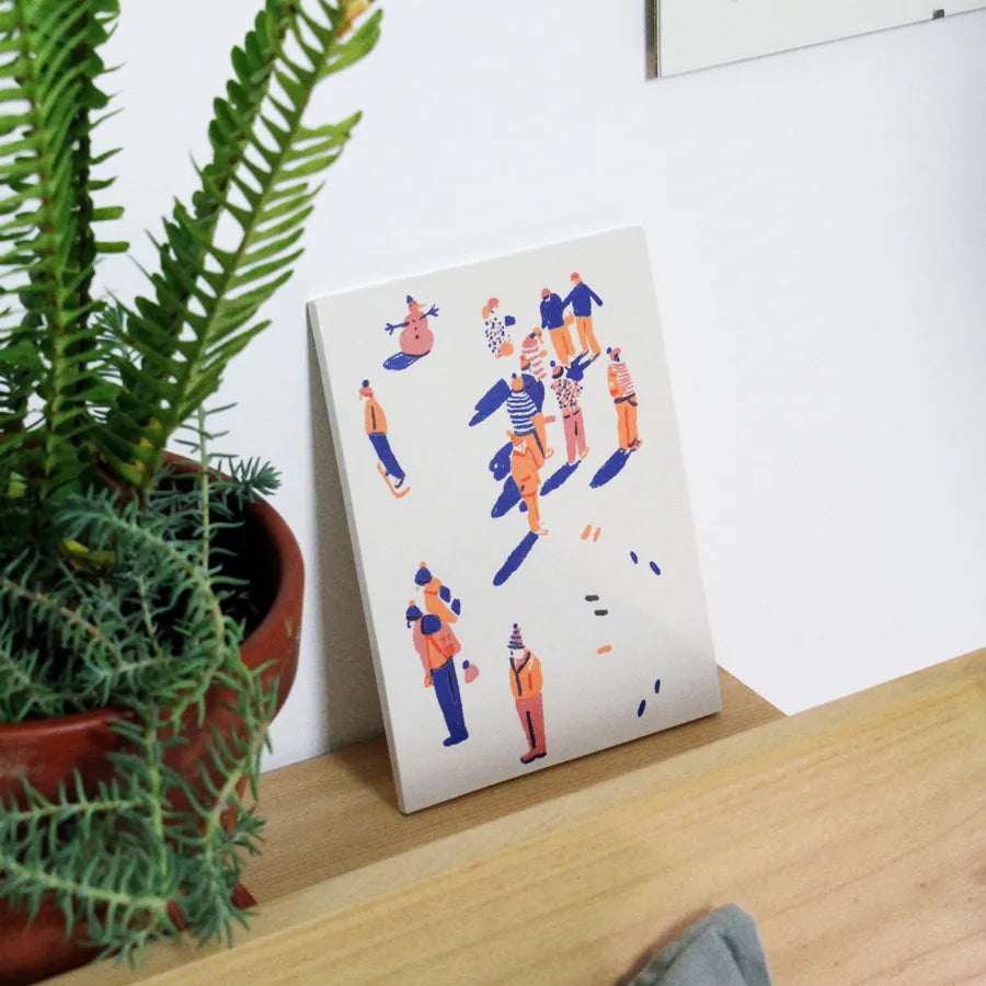 screen-printed tile with colorful illustrations of people on the snow on a wooden surface next to a potted plant.