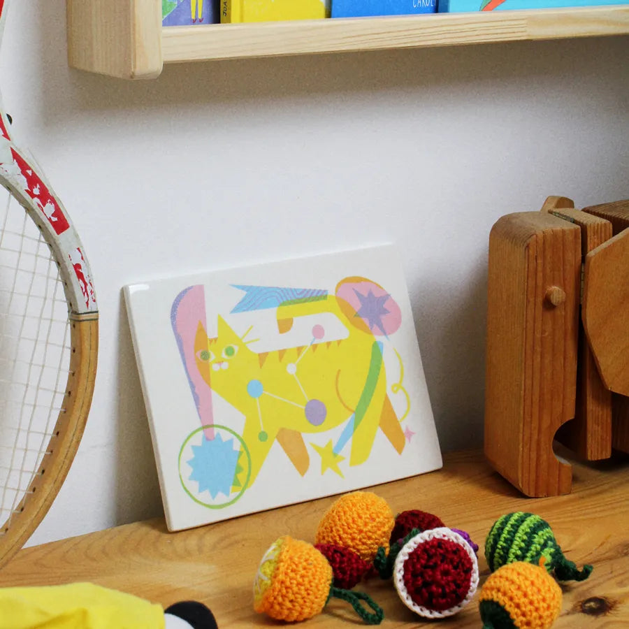 Colorful cat illustration on a handmade tile with yarn balls and a tennis racket in the background.