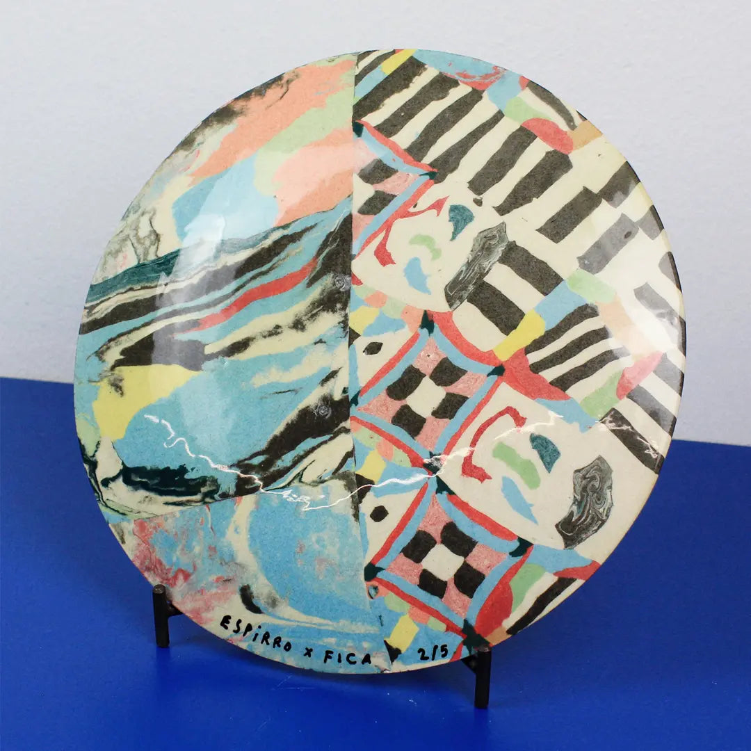 A round ceramic plate with a split abstract design, displayed on a small black stand against a blue and gray background. The left half features fluid marbled patterns in blue, yellow, pink, and black, while the right half features a geometric checkered pattern in black, white, red, and blue.