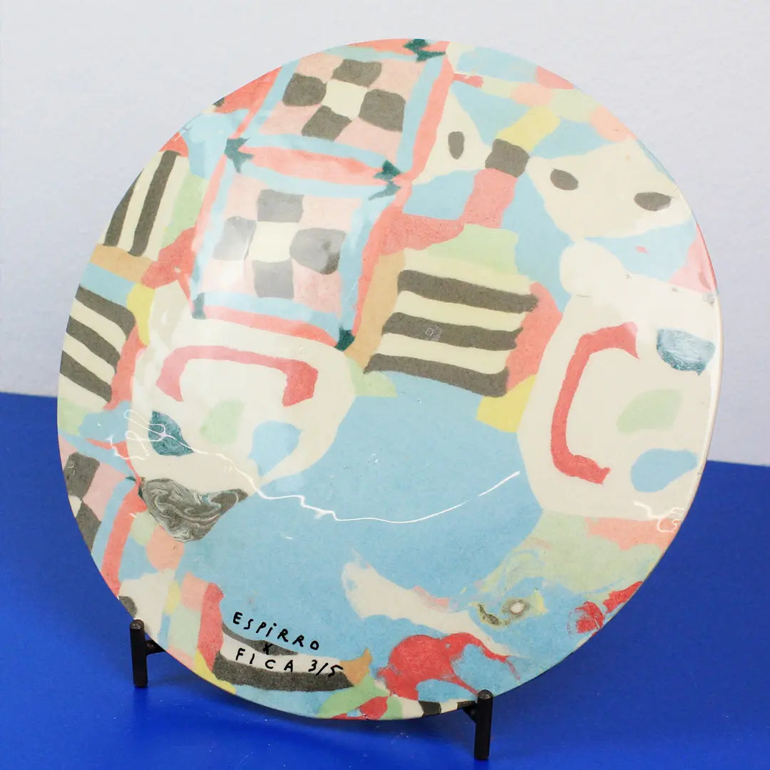 A round ceramic plate with an abstract hand-painted design in pastel blue, pink, red, green, yellow, and black, displayed on a small black stand against a light gray background and blue surface.