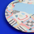 A close-up of a round ceramic plate featuring a colorful abstract pattern with a light blue center, white background, and hand-painted lines in pink, red, green, yellow, black, and gray, set against a solid blue background.
