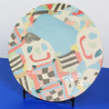 A round ceramic plate with a colorful abstract hand-painted pattern, displayed on a small black stand against a split white and blue background.