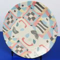 A round ceramic plate with a colorful abstract geometric pattern, displayed on a small black stand against a split white and blue background.
