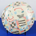 A round ceramic plate with a colorful abstract hand-painted pattern, displayed on a small black stand against a blue and gray background. The plate features a cream base with pink, light blue, yellow, gray, and black geometric and organic shapes.