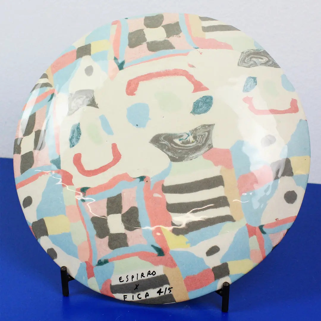 A round ceramic plate with a colorful abstract hand-painted pattern, displayed on a small black stand against a blue and gray background. The plate features a cream base with pink, light blue, yellow, gray, and black geometric and organic shapes.